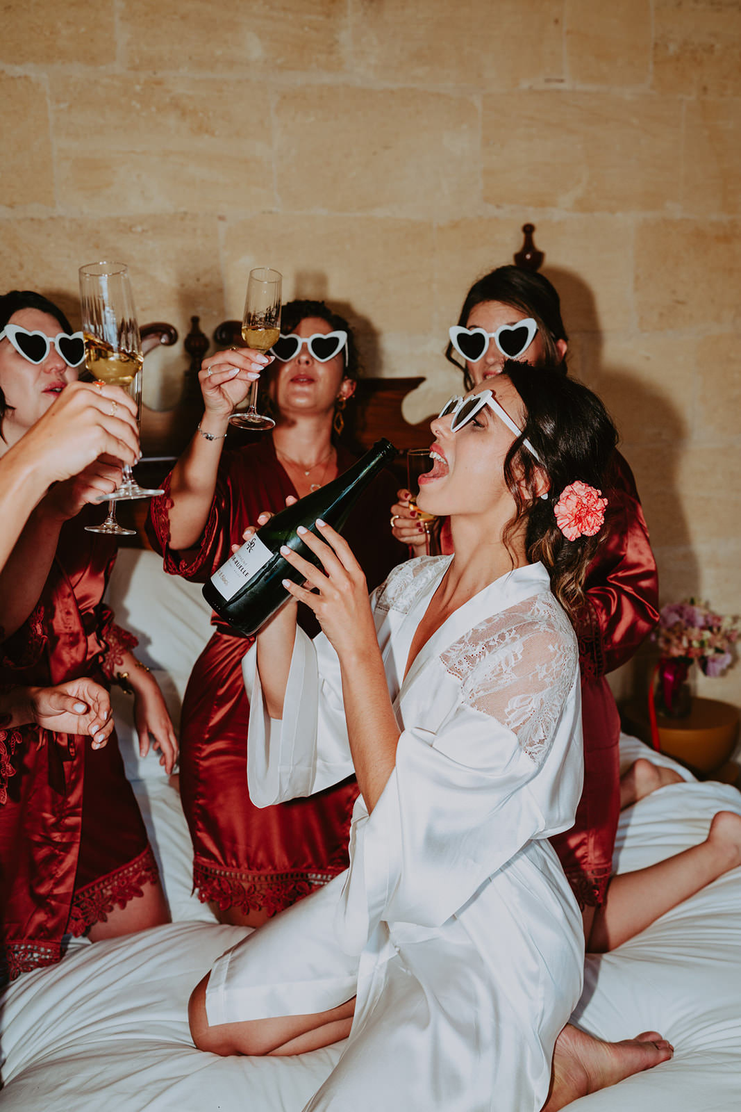 photo d'une marié sur un lit avec ses copines qui boit joyeusement au goulot une bouteille de champagne
